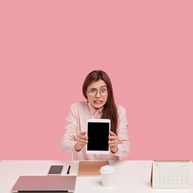 Foto der verwirrten jungen Frau hält Touchpad mit Mock-up-Bildschirm, verwendet Anwendung, trägt transparente Brille