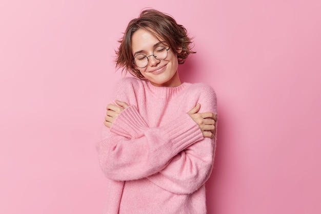 Foto der zarten hübschen frau mit kurzen haaren umarmt sich selbst fühlt sich in einem neuen weichen pullover wohl und trägt eine runde brille zur sehkorrektur, die über rosafarbenem hintergrund isoliert ist selbstpflegekonzept