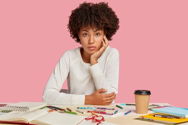 Foto des attraktiven schwarzen Designers mit Afro-Haarschnitt, sitzt am Arbeitsplatz, trägt weißen Pullover, macht Zeichnungen mit Buntstiften, isoliert über rosa Wand, genießt Kaffee