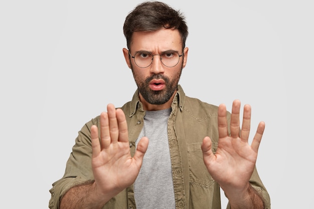 Foto des bärtigen jungen Mannes zeigt Stoppgeste, hat unzufriedenen Gesichtsausdruck, bestreitet etwas, spricht über verbotene Dinge, trägt modisches Hemd, isoliert über weißer Wand