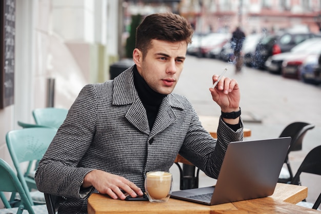 Foto des eleganten Geschäftsmannes mit dem Brütenblick, der draußen im Café sitzt, Zigarette raucht und Cappuccino trinkt