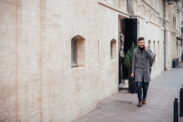 Foto des eleganten Mannes im Mantel mit Tasche in der Hand gehend hinunter leere Straße und auf Smartphone sprechend