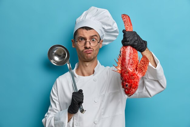 Foto des ernsthaften professionellen Kochs hält Schöpflöffel und Fisch, bereitet Gourmet-Meeresfrüchtegericht zu, trägt weiße Uniform, schwarze Handschuhe, kocht Suppe des roten Basses