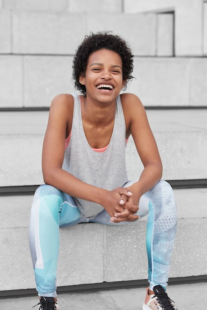 Foto des fröhlichen dunkelhäutigen Jungen mit den knackigen schwarzen Haaren, lächelt breit, gekleidet in lässiger Weste und in den Leggings, sitzt auf Stufen, genießt gesunden Lebensstil, Lieblingssportspiel. Vitalität, Wellness