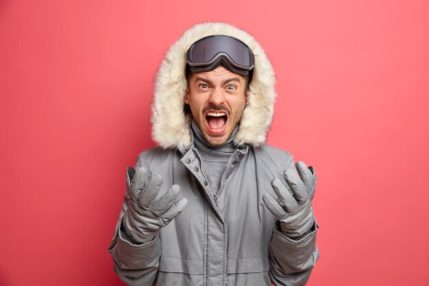 Foto des gereizten europäischen Mannes schreit laut und Gesten drücken wütend negative Gefühle aus, trägt warme Thermojackenhandschuhe.