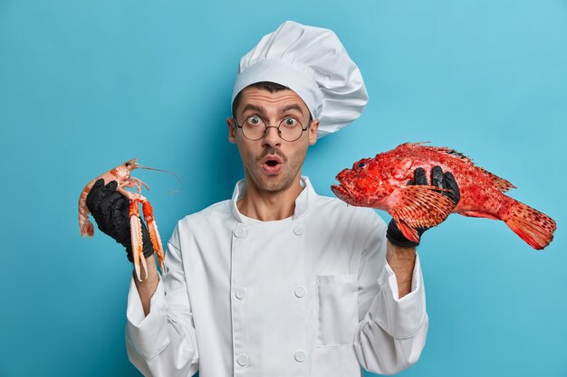 Foto des geschockten erfahrenen Küchenchefs hält Krebse, Rotbarsch, kocht köstliches Gericht aus Meeresfrüchten, arbeitet an seiner Fischkarte, hält den Mund vor Staunen offen