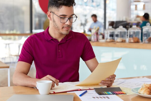 Foto des hübschen dunkelhaarigen jungen Mannes hält Dokument, liest Informationen, hat Notizbuch geöffnet, studiert Grafik und Diagramme, trägt Freizeitkleidung, trinkt aromatischen Kaffee, posiert in gemütlichem Café