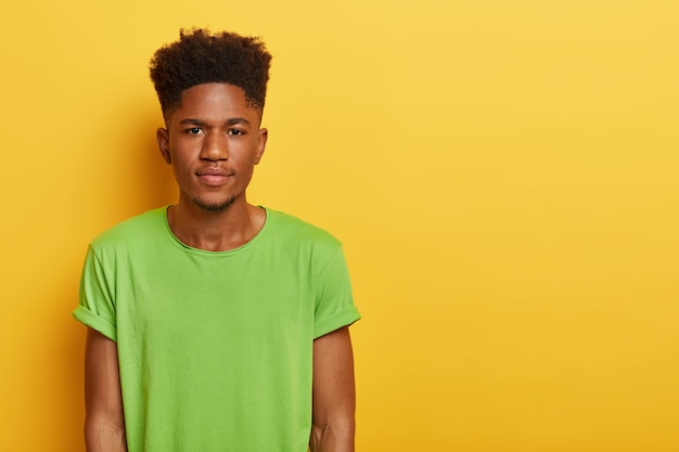 Foto des hübschen Teenagers mit dunkler Haut, lockiger Frisur, trägt lässiges grünes T-Shirt, sieht mit ruhigem ernstem Ausdruck aus