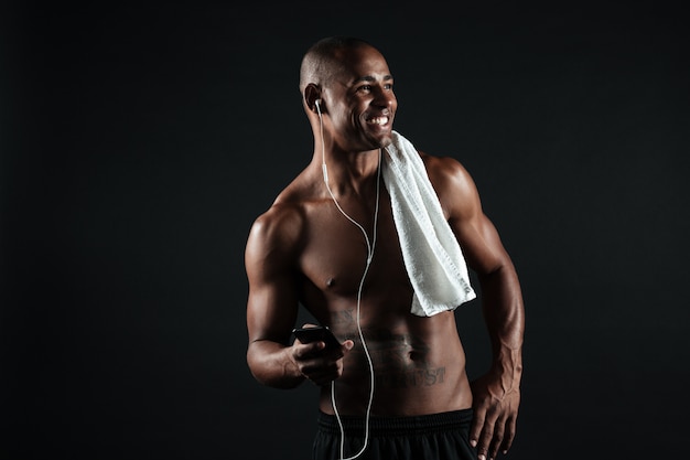 Foto des jungen lächelnden afroamerikanischen Sportmannes mit Handtuch, der Telefon hält und Musik hört