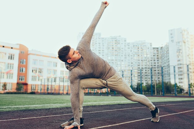 Foto des jungen Mannes in voller Länge beim Training am Morgen im Stadion. Er trägt einen grauen Sportanzug. Er macht Sport.