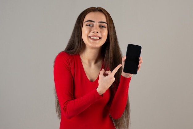 Foto des lächelnden entzückenden Mädchens im roten Sweatshirt, das Mobiltelefon auf grauer Wand hält.