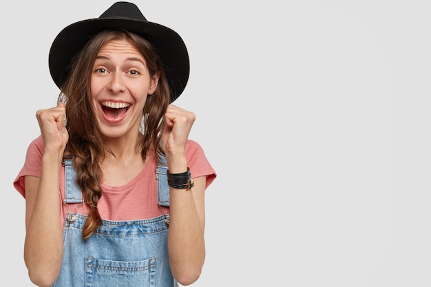 Foto des Landarbeiters im stilvollen Hut lässiges T-Shirt und Overalls, ballt die Fäuste mit Triumph, erreicht schließlich perfektes Ergebnis, fühlt sich vom Sieg erstaunt, isoliert über weiße Wand, freier Raum