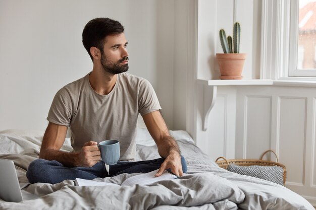 Foto des ruhigen gutaussehenden unrasierten Mannes liest gern Bestseller, hält Tasse mit Kaffee oder Tee, sitzt gekreuzte Beine im Bett, denkt über Lebenssituation nach, schaut nachdenklich zur Seite. Menschen- und Hobbykonzept