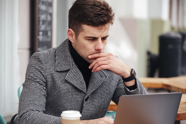 Foto des sachlichen Mannes arbeitend mit silbernem Laptop im Café draußen, trinkender Mitnehmerkaffee