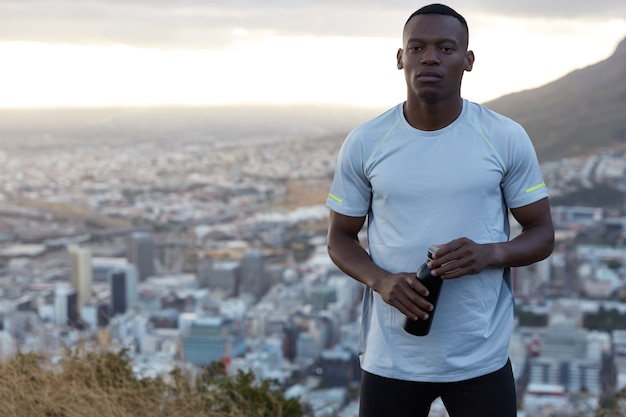 Foto des sportlichen muskulösen schwarzen Mannes im lässigen T-Shirt, trainiert am frühen Morgen, trägt Flasche mit Getränk, ist in guter körperlicher Form, steht auf Hügel über verschwommener Großstadt, freier Raum