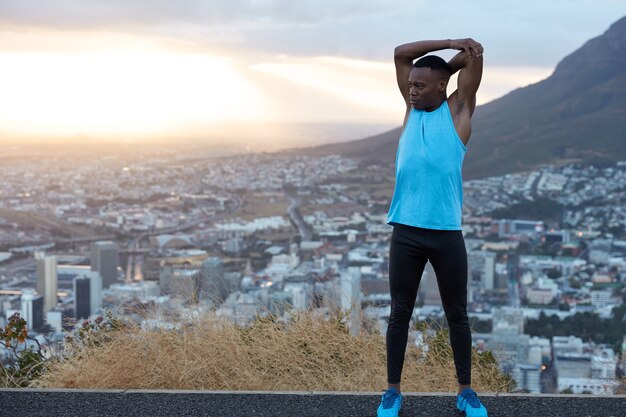 Foto des starken Mannes mit guter Flexibilität hebt Hände über Kopf, macht Sportübungen unter freiem Himmel auf Hügel gegen Panoramablick mit Sonnenaufgang, Stadtgebäude und Felsen. Sportlicher Schwarzer hat Training