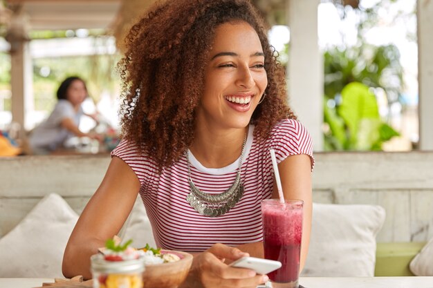 Foto des überglücklichen dunkelhäutigen Mädchens hat buschiges Haar, lacht und schaut zur Seite, benutzt Handy für Online-Kommunikation und Nachrichten mit Freunden, trinkt Smoothie in der gemütlichen Cafeteria