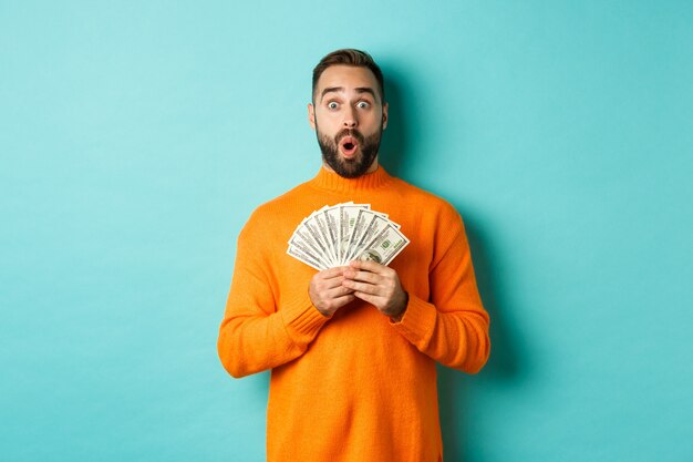 Foto des überraschten Kerls, der Geld hält, erstaunt aussieht und mit Dollars gegen türkisfarbene Wand steht