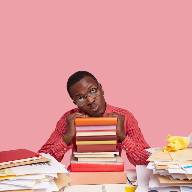 Foto des unzufriedenen afroamerikanischen Studenten mit mürrischem Ausdruck, hält Hände auf Stapel von Lehrbüchern, neigt Kopf, gekleidet in rosa Hemd