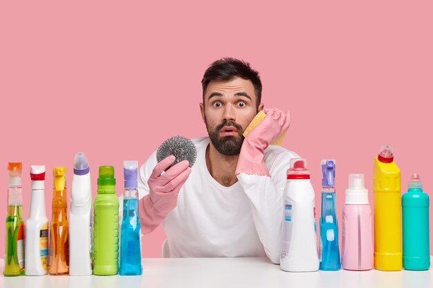 Foto des unzufriedenen bärtigen Mannes in Gummihandschuhen hält Lappen zum Reinigen des Küchenherds, sieht verlegen aus