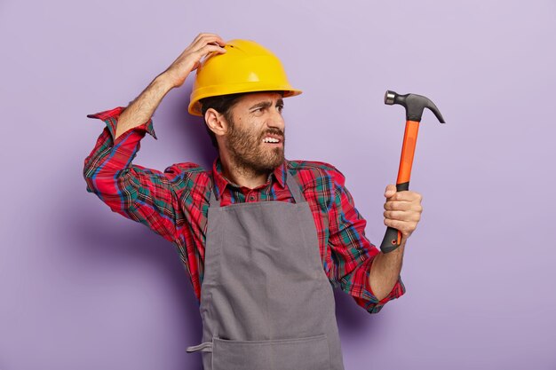 Foto des unzufriedenen Handwerkers biegt nach rechts ab, schaut mit gerunzelter Stirn in die Ferne, hält Hammer, ist professioneller Baumeister, bemerkt neues Objekt zur Reparatur trägt Helm, Schürze. Renovierung, Technik