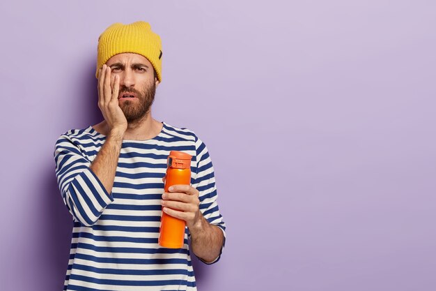 Foto des unzufriedenen Mannes hält Hand auf Wange, trägt Flasche mit Kaffee oder Tee, hat schläfrigen müden Ausdruck, trägt Freizeitkleidung