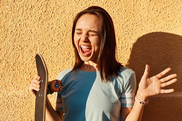 Foto einer aufgeregten dunkelhaarigen Frau mit blauem T-Shirt, die gegen die gelbe Wand im Freien steht und glücklich schreit, Longboard in den Händen hält und Glück ausdrückt.