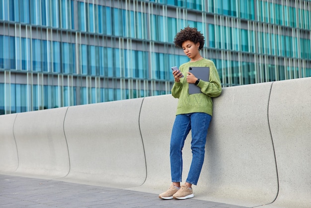 Foto einer ernsten jungen lockigen Frau steht in voller Länge vor urbanem Hintergrund verwendet moderne Smartphone- und Tablet-Checks Social Newsfeed erhielt Benachrichtigung hat einen eigenen Blog trägt stilvolle Kleidung