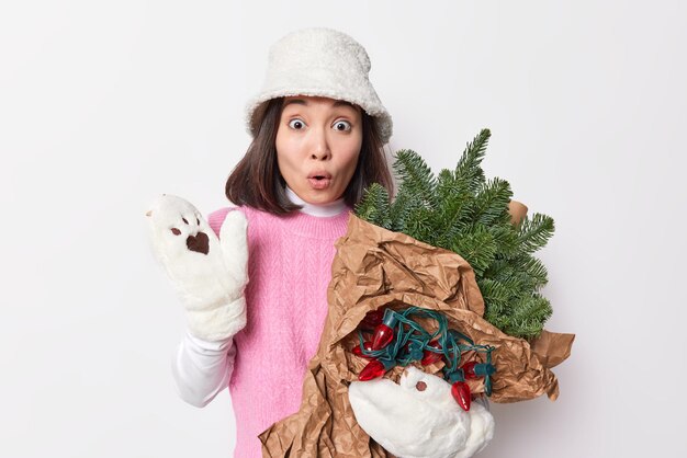 Foto einer fassungslosen Frau, die den Atem anhält, fühlt sich betäubt, trägt Winterkleidung, trägt immergrüne Fichtenzweige, die in Papier gewickelt sind und Girlanden kehrt vom Weihnachtsmarkt isoliert über weiße Wand zurück