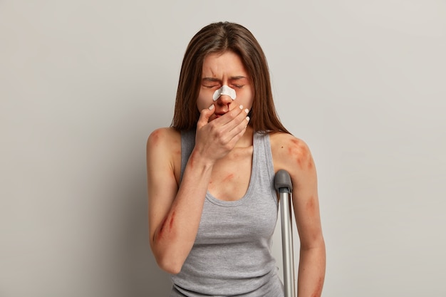 Foto einer Frau, die von jemandem geschlagen wurde, Opfer von Gewalt oder Vergewaltigung ist, eine blutende Nase, viele blaue Flecken am Körper und Brüche hat, die Hand im Gesicht hält, die Augen vor Schmerzen schließt und mit der Krücke im Haus steht