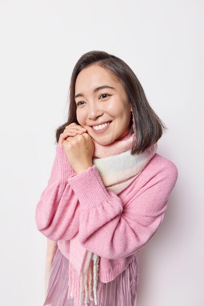 Foto einer gutaussehenden asiatischen Frau, die sanft lächelt, hält die Hände in der Nähe des Gesichts, hat einen verträumten Ausdruck, denkt an etwas Angenehmes, trägt einen rosa Pullover und einen Schal um den Hals, isoliert auf weißem Hintergrund