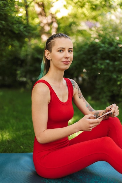 Foto einer schönen Frau in rotem Overall, die mit Handy auf einer Yogamatte sitzt und verträumt in die Kamera schaut, während sie Yoga im Park praktiziert