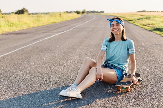 Foto einer schönen lächelnden Frau, die T-Shirt, Haarband und kurze Haare trägt, mit Longboard auf der Straße sitzt, die Kamera mit einem zahnigen Lächeln betrachtet, Glück ausdrückt, Freizeit genießt.