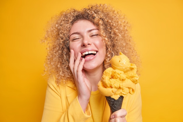 Foto einer überglücklichen, lockigen Frau, die sich amüsiert lächelt, hält die Augen geschlossen und genießt es, köstliches Eis mit Mangogeschmack zu essen, mit leckerem gefrorenem Dessert einzeln auf gelber Wand.