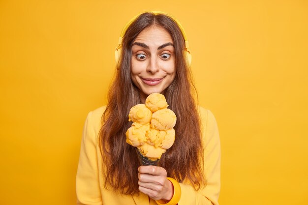 Foto einer überraschten, fröhlichen europäischen Frau mit langen dunklen Haaren starrt auf appetitliches Eis, das verhungert, um es zu essen, hört Musik über drahtlose Stereo-Kopfhörer posiert drinnen gegen gelbe Wand