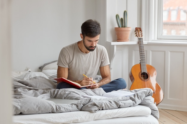 Foto eines ernsten Mannes mit dunklen Borsten, schreibt eine Geschichte in ein Tagebuch darüber, was tagsüber passiert ist, sitzt mit gekreuzten Beinen im Bett, trägt Freizeitkleidung, hat freien Tag oder ein Wochenende. Männliche Aufzeichnungen Informationen