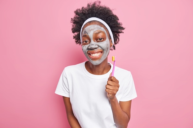 Foto eines fröhlichen tausendjährigen Mädchens mit Afro-Haar, das angenehm lächelt, trägt Tonmaske auf und hält Zahnbürste