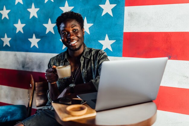 Foto eines glücklichen afrikanischen Mannes, der in einem Café sitzt und am Laptop arbeitet, eine Kaffeepause einlegen.