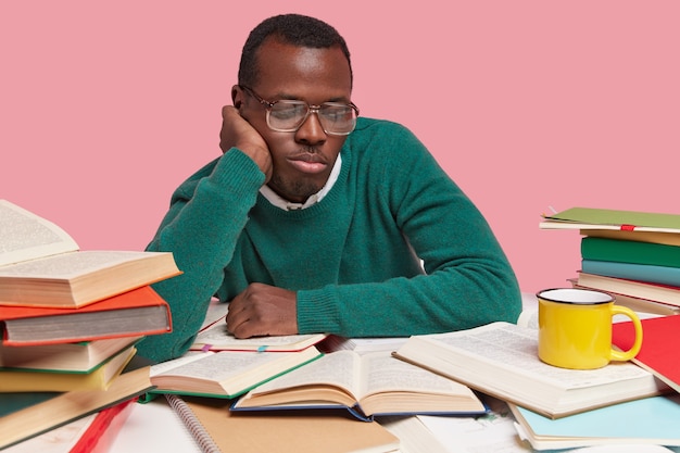 Foto eines konzentrierten schwarzen Mannes, der in geöffneten Büchern fokussiert ist, am Lesen beteiligt ist, einen grünen Pullover trägt und neue Informationen herausfindet