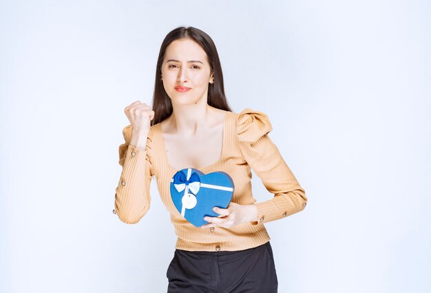 Foto eines Modells der jungen Frau, das eine herzförmige Geschenkbox gegen weiße Wand hält.