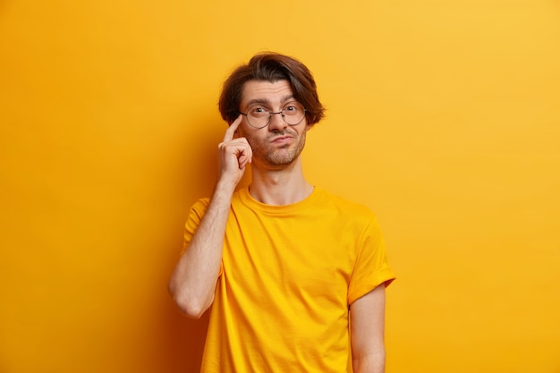 Foto eines nachdenklichen jungen Europäers hält den Finger auf den Tempel und stellt sich vor, dass etwas eine runde Brille trägt und ein lässiges T-Shirt, das über einer gelben Wand isoliert ist, eine wichtige Entscheidungsvariante ist