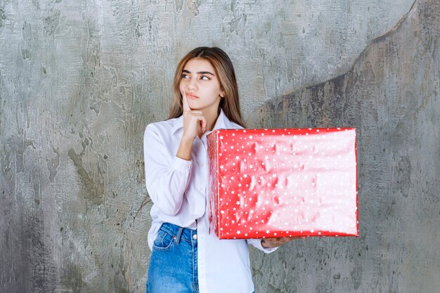 Foto eines nachdenklichen Mädchenmodells mit langen Haaren, das ein großes rotes Geschenk hält