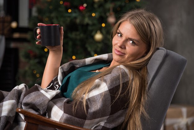 Foto eines ruhigen Mädchens, das in einer karierten karierten Decke sitzt und eine Tasse mit heißem Getränk hält