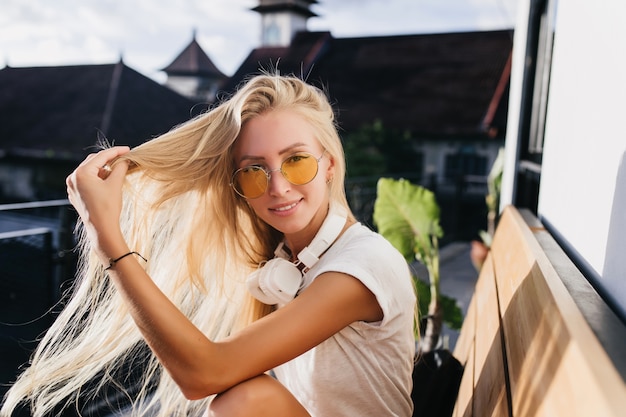 Foto im Freien der siegreichen Dame in der gelben Sonnenbrille, die mit ihren langen blonden Haaren auf Stadthintergrund spielt.