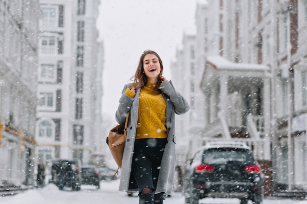 Foto im Freien des anmutigen weiblichen Modells im trendigen langen Mantel, der in der Stadt am Wintertag entspannt. Gut aussehende junge Frau im gelben Pullover, die Spaß beim Einkaufen im Dezembermorgen hat.