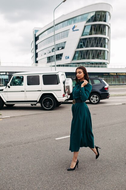 Foto in voller Länge einer hübschen brünetten Frau in einem langen smaragdgrünen Kleid mit Knöpfen und schwarzen Lederabsätzen, die selbstbewusst die Straße entlang gegen moderne Gebäude und Autos laufen.