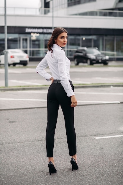 Foto in voller Länge einer stilvollen Frau in schwarzer Hose und weißem Hemd, die auf der Straße gegen das moderne Gebäude steht. Stil- und Modekonzept