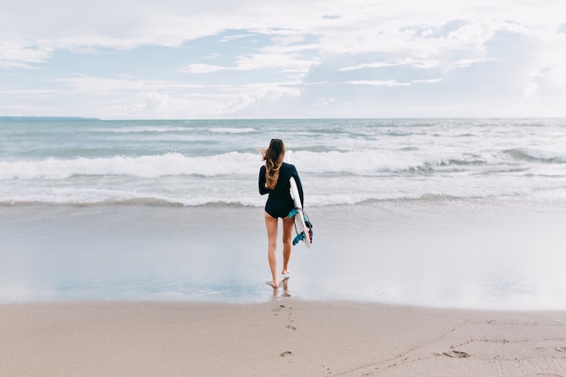 Foto in voller Länge von der Rückseite einer attraktiven jungen Frau mit langen Haaren im Badeanzug läuft im Meer mit einem Surfbrett, Hintergrund des Ozeans, Sport, aktiver Lebensstil