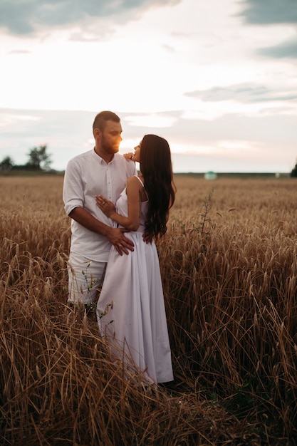 Foto in voller Länge von einem romantischen Paar in weißer Kleidung, das sich bei Sonnenuntergang im Weizenfeld umarmt.