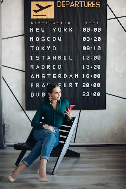 Foto in voller Länge von eleganter Geschäftsfrau in Smart Casual mit Handy, die auf ihren Flug gegen Abflugplakat wartet.